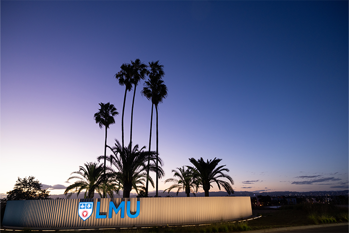 LMU sign lit up in front of palm tree with dusk sky and city behind it